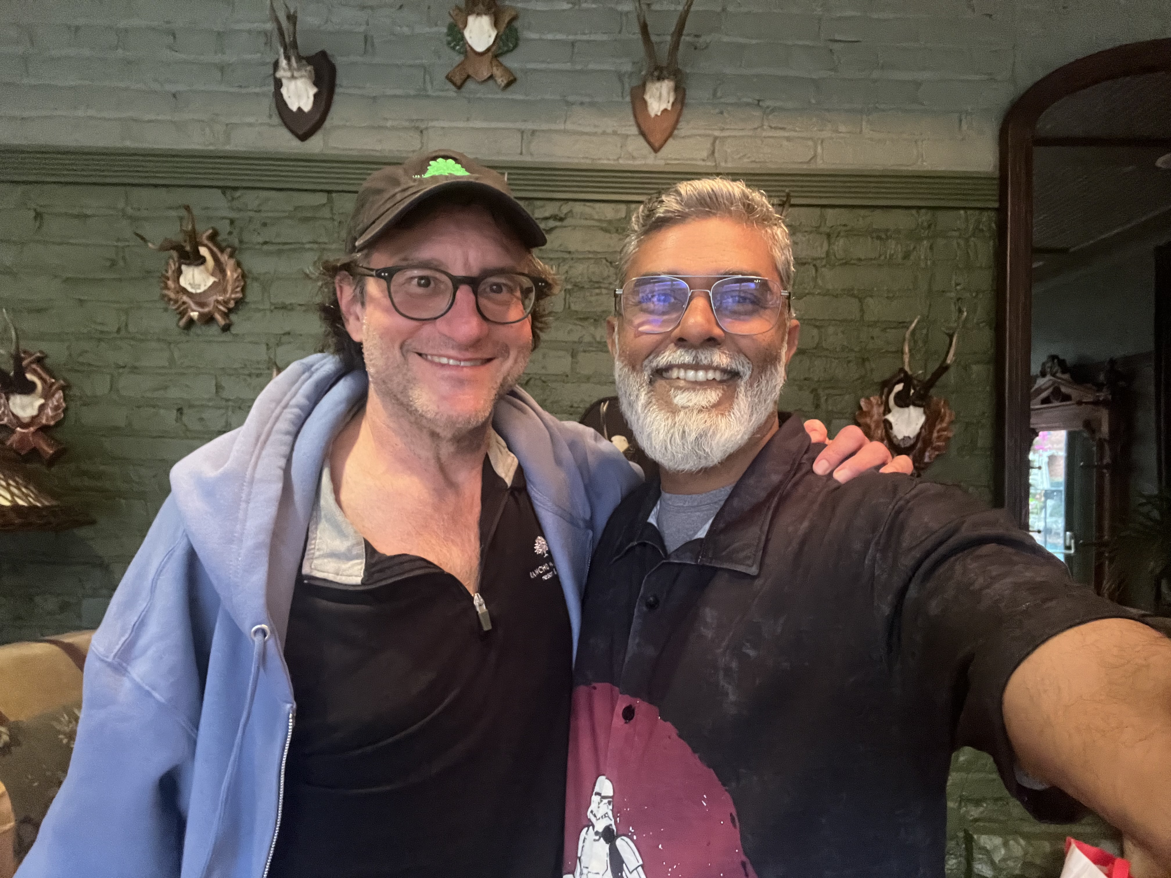Two smiling men posing for a selfie indoors, with decorative wall mounts in the background.