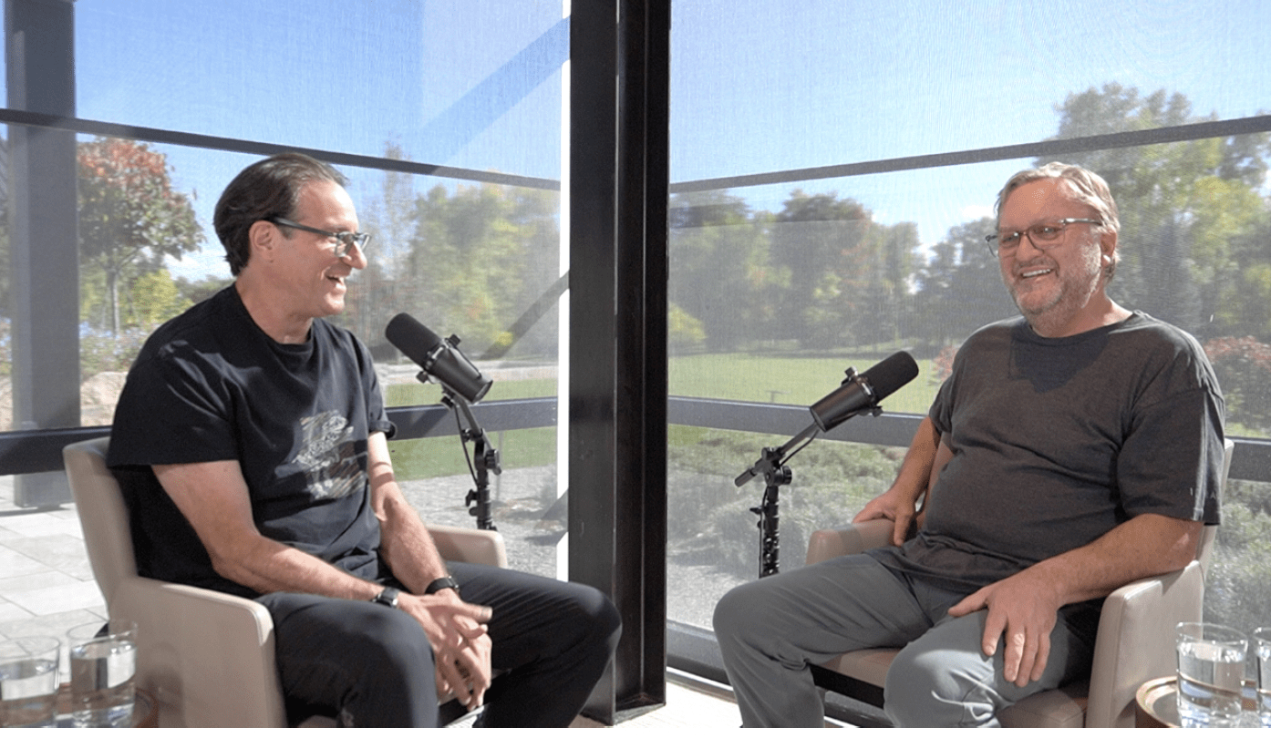 Two men sitting in chairs in a bright, modern setting, engaged in conversation with microphones in front of them.